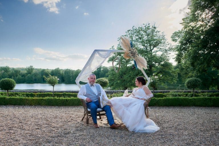 Photo fun d'un couple de mariés dont la mariée à poser ses jambes chaussées de baskets sur les cuisses de son mari. En arrière le plan, la rivière passant dans le domaine du Château de Verrerie à Oizon.